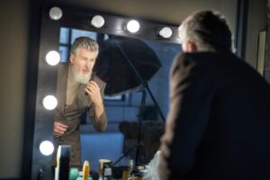 Attraktiver grauhaariger Mann mittleren Alters im eleganten Anzug, der sich im Spiegel betrachtet, während er sich auf ein Fotoshooting im Studio vorbereitet